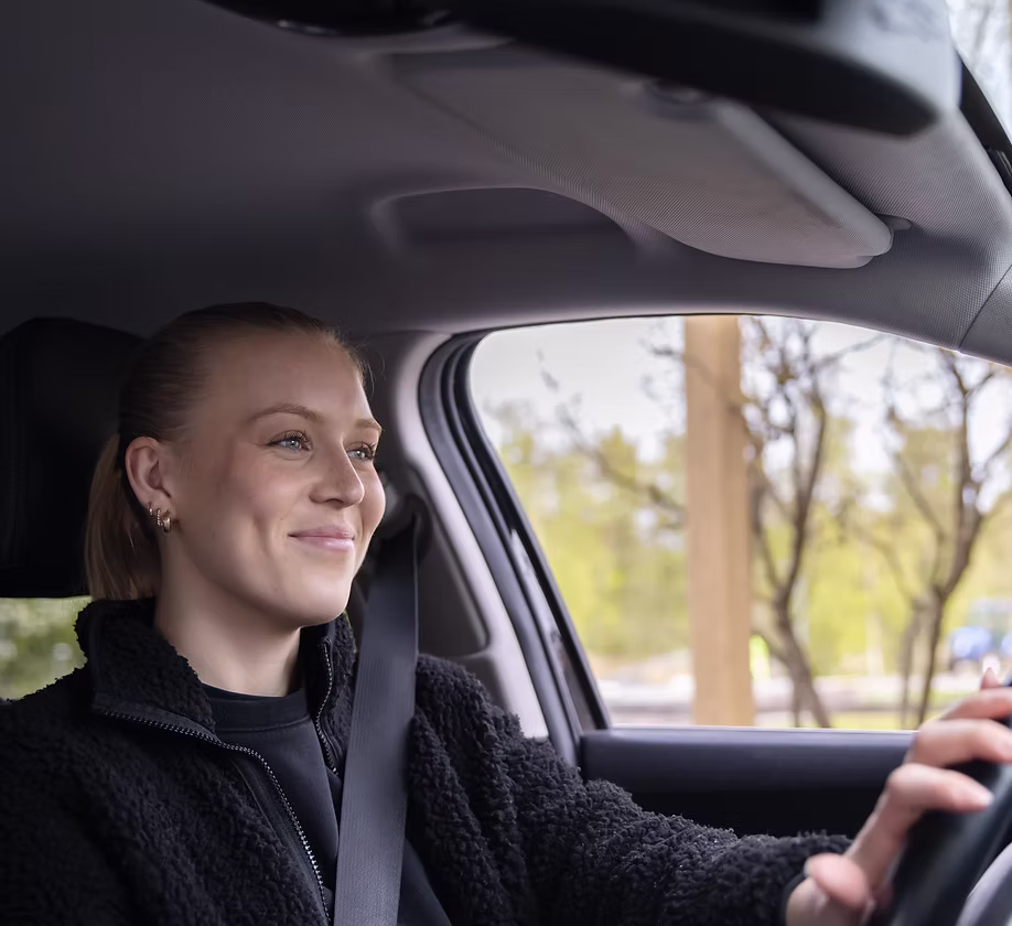 Leende WeDrive-förare i arbetskläder bakom ratten med bilbältet på, vårgrönska genom sidorutan